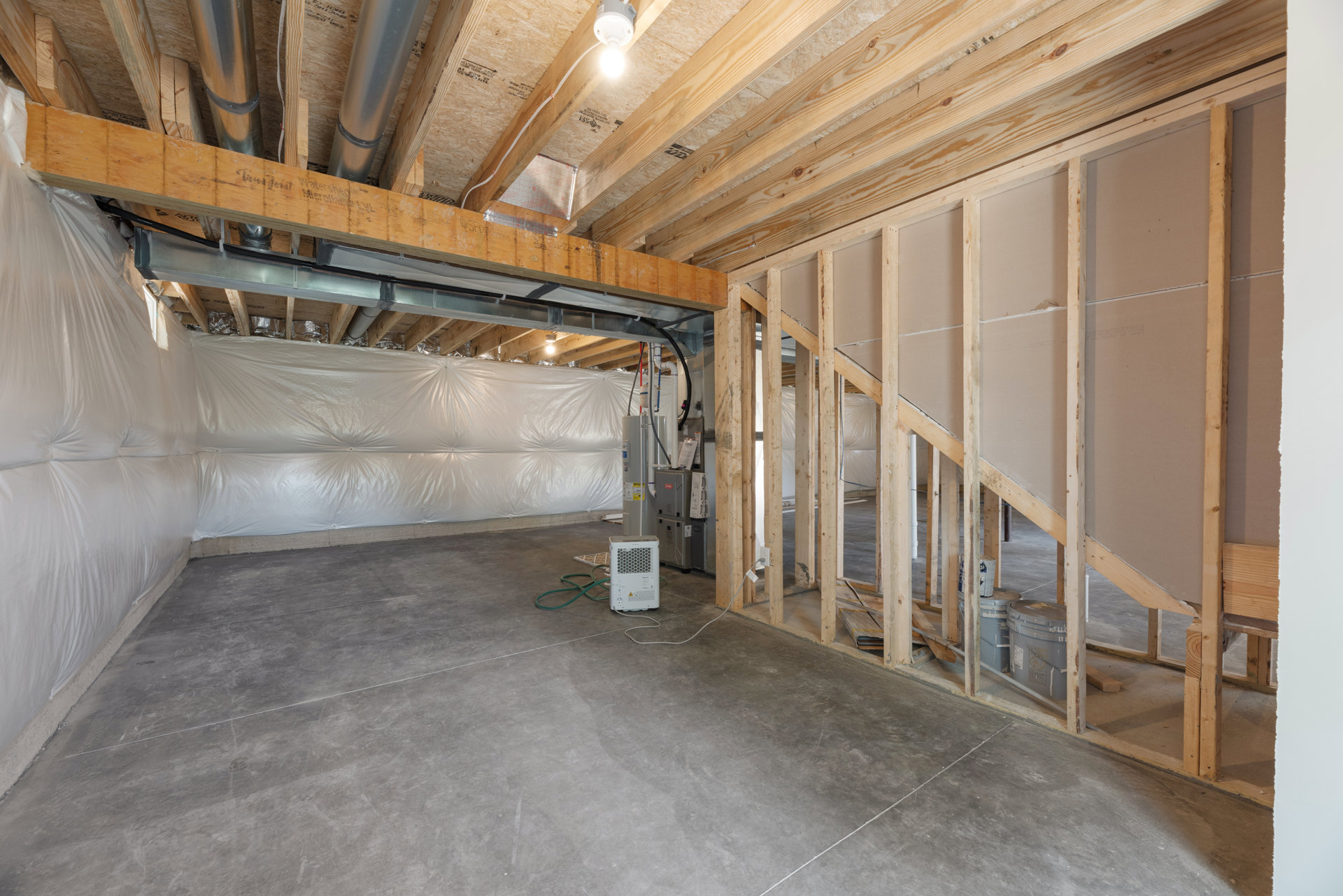 Exposed wood ceiling beams, white plaster walls, ceiling-mounted light bulb, metal box, and white plastic insulation covering in a basement room.