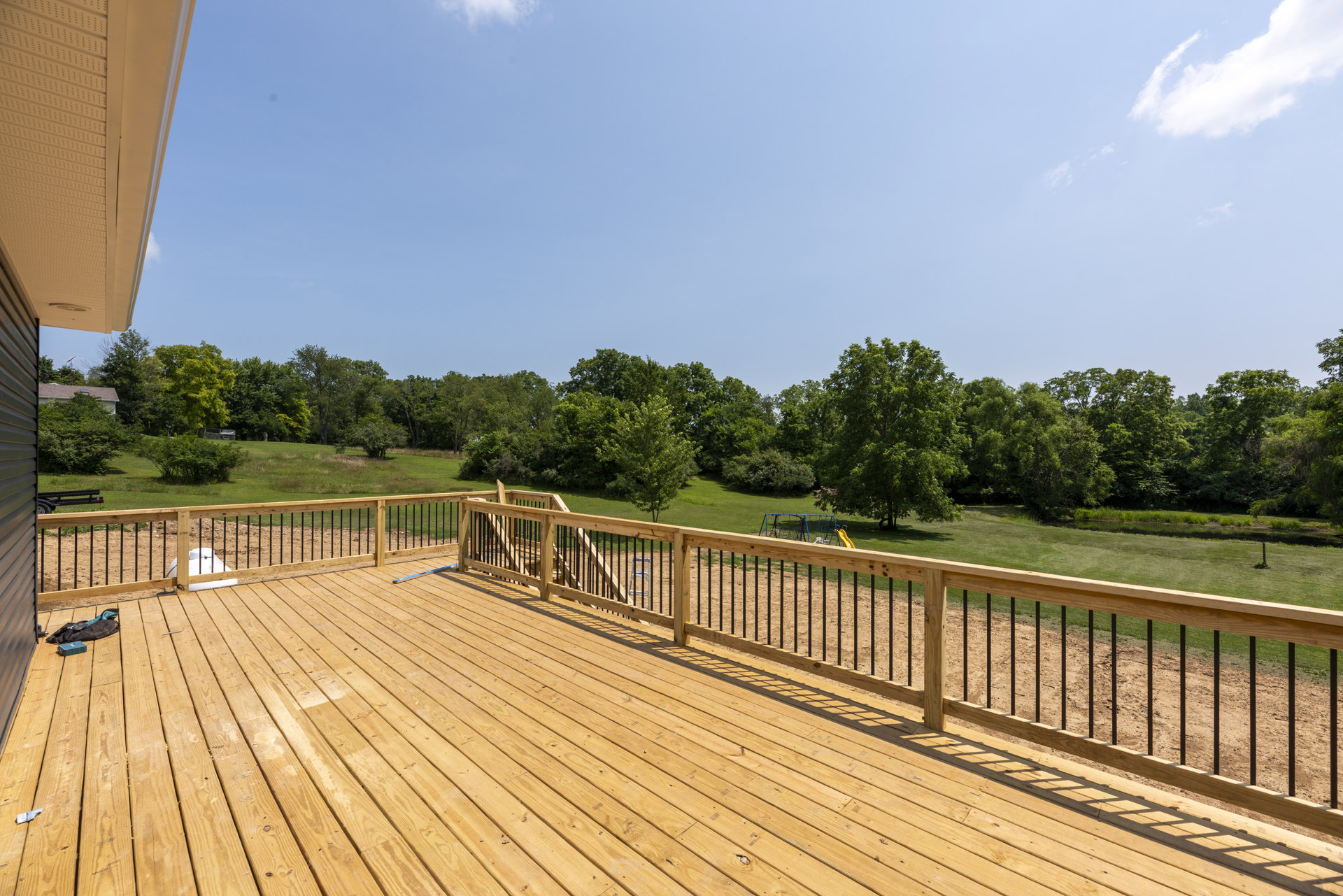 Wooden deck with horizontal plank flooring, fenced perimeter, mature trees and blue sky in the background, tool bag resting on deck, playground equipment partially visible beyond