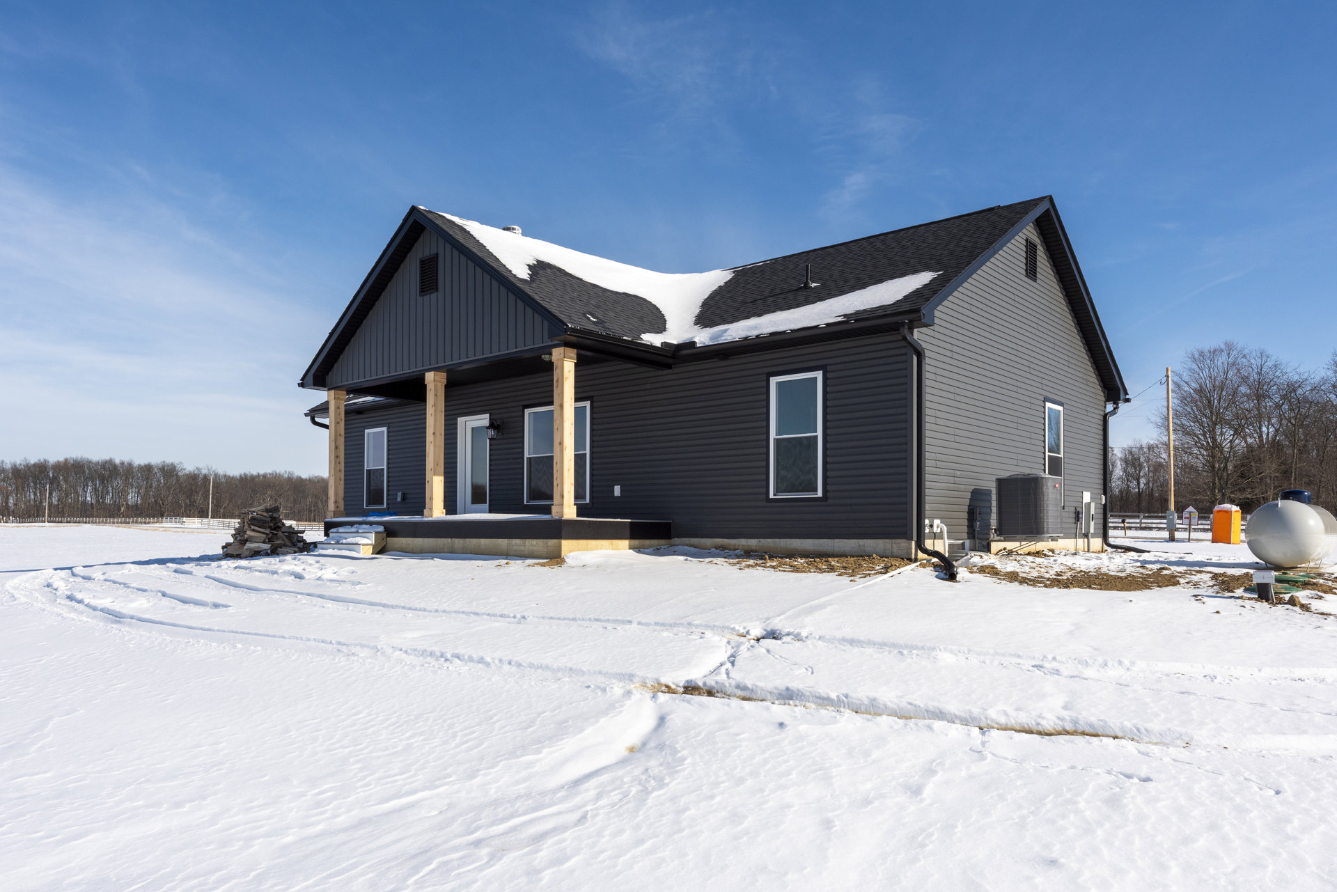 Two-story house with white-framed windows, snow covering the roof and ground, visible tracks in snow, wooden post near window, large white round object with blue cap in yard, bare
