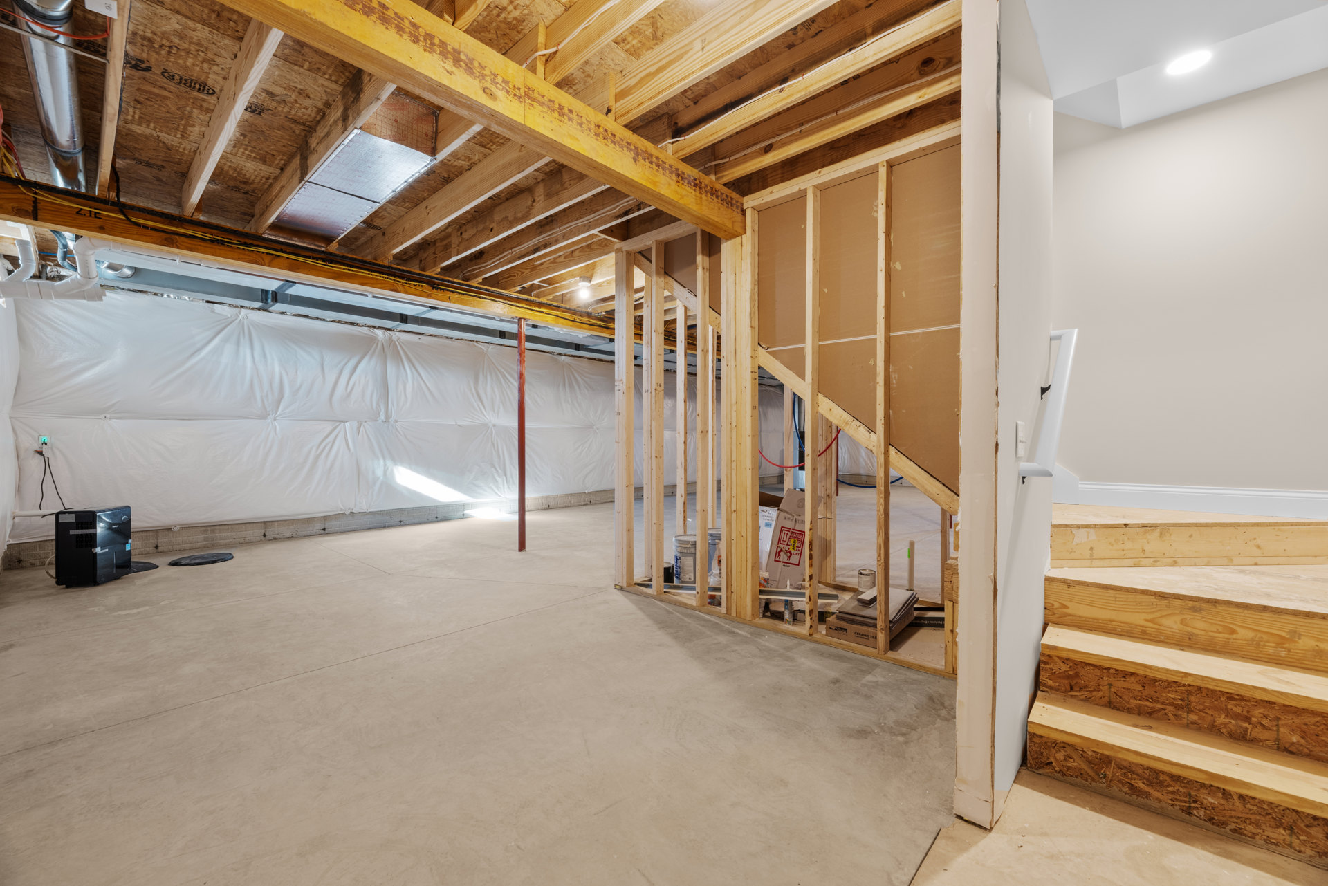 Open-concept room featuring unfinished wood staircase, exposed beams, white plaster walls, and construction materials including a computer tower and a white sheet draped along one