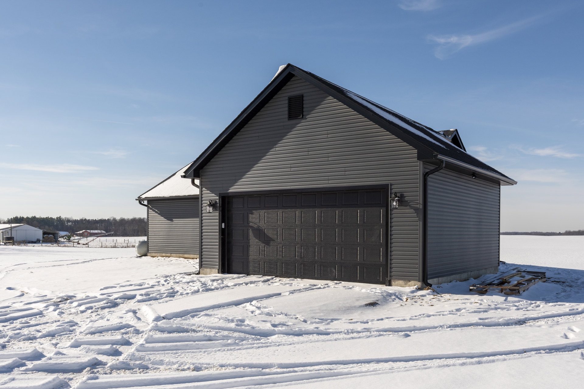Modern house with black garage door, exterior light fixture, snow-covered driveway with tire tracks, white siding, and snowy landscape.