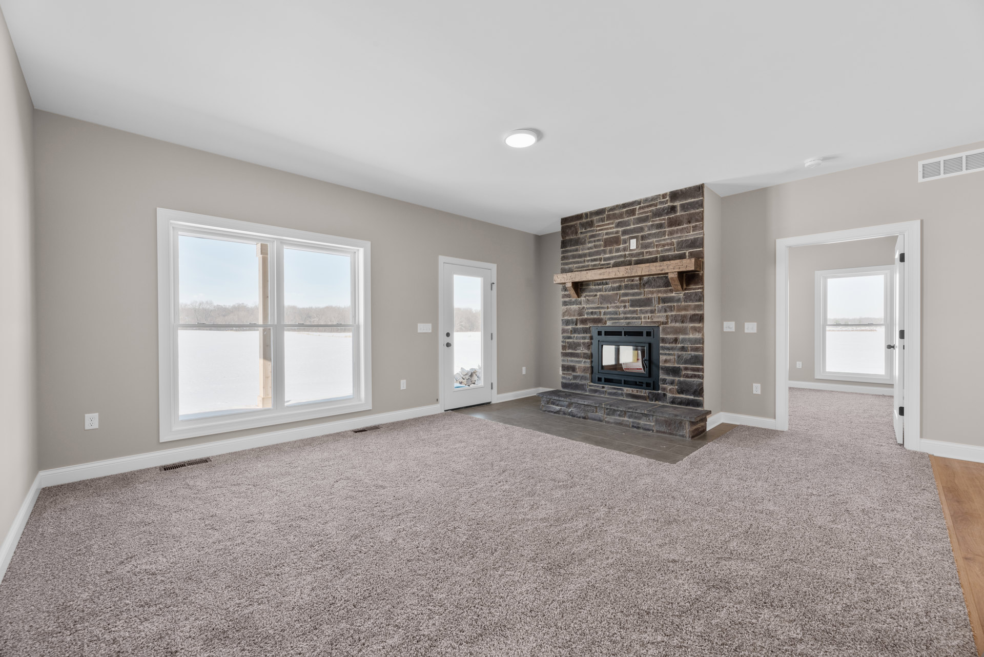 Living room with beige carpet, central fireplace featuring black surround and glass window, large window and door revealing snowy landscape, neutral walls and ceiling