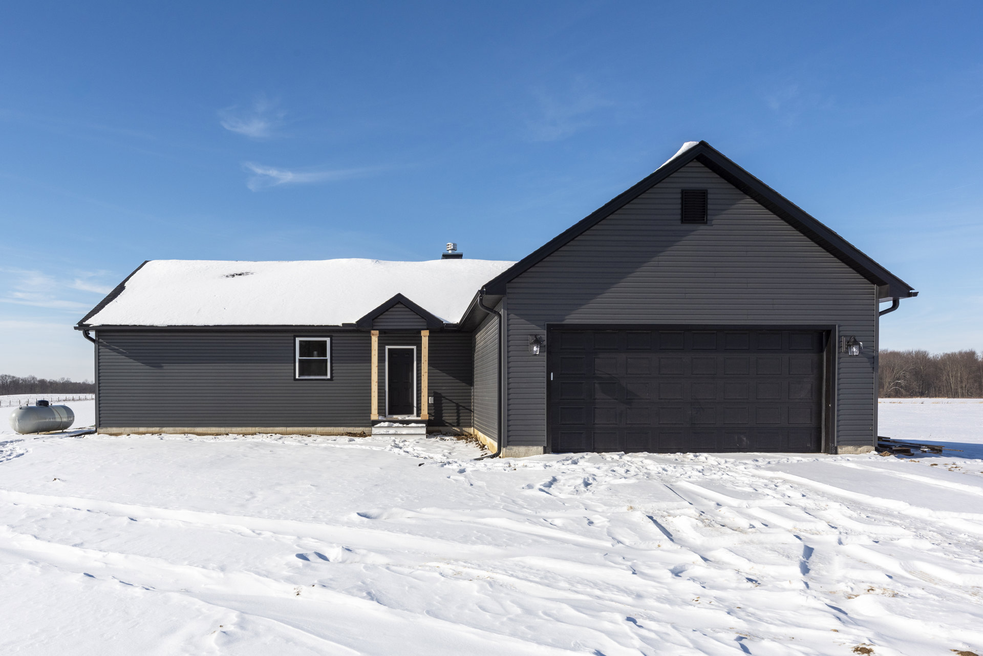 Two-story cottage-style home with white siding, black-framed windows, attached garage, black front door, large white propane tank beside the house, snow covering the ground and