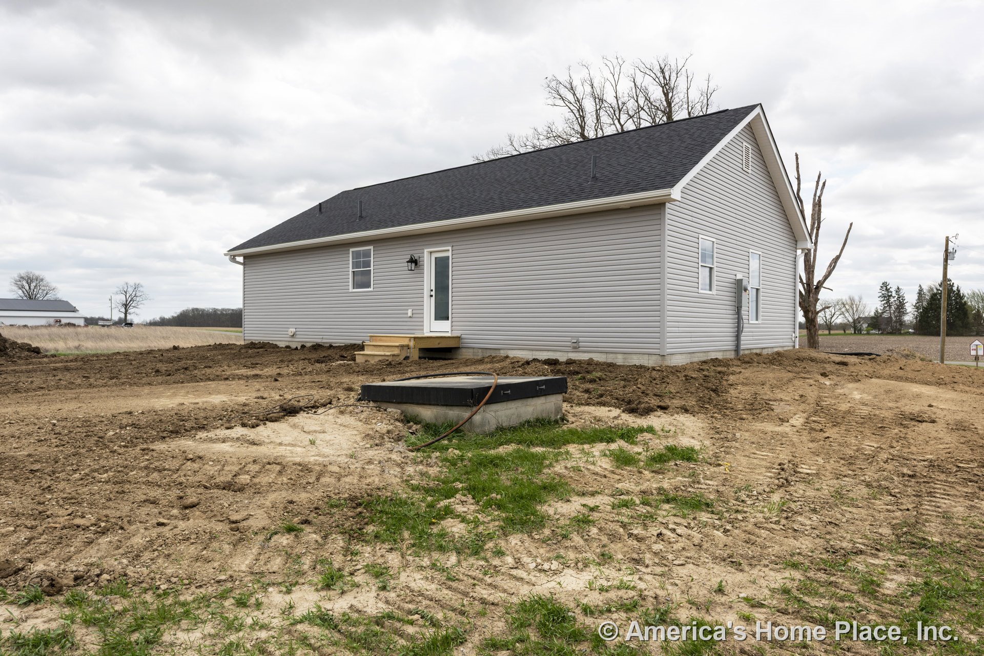 Gray vinyl siding single-story home with black shingle roof, white trim, double-hung windows, rear glass entry door, small wooden steps, utility meter, and outdoor lighting.
