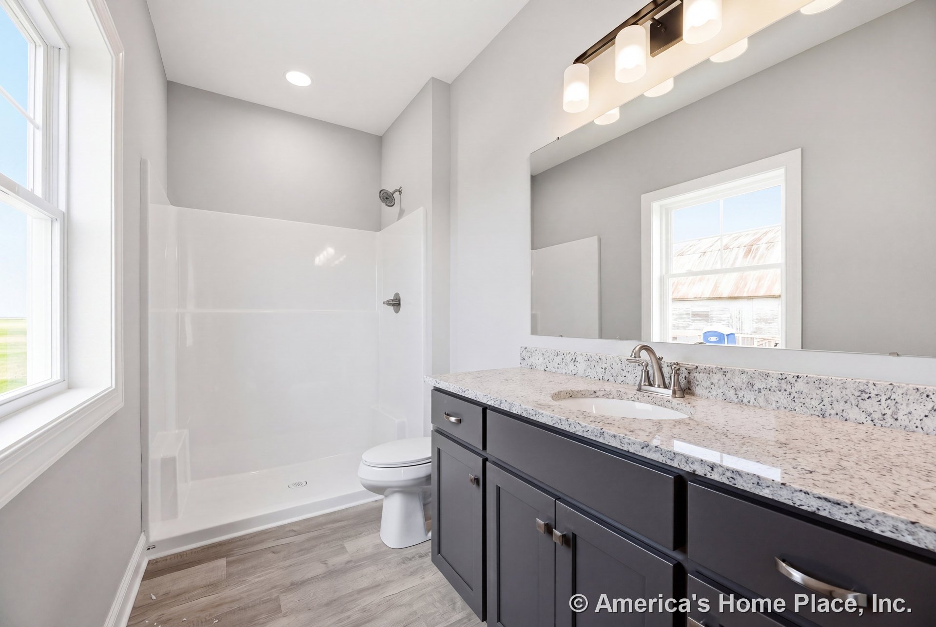 Walk-in shower enclosure beside granite vanity countertop with dark shaker cabinets, large wall mirror, recessed ceiling light, window framed in white trim, single sink, modern