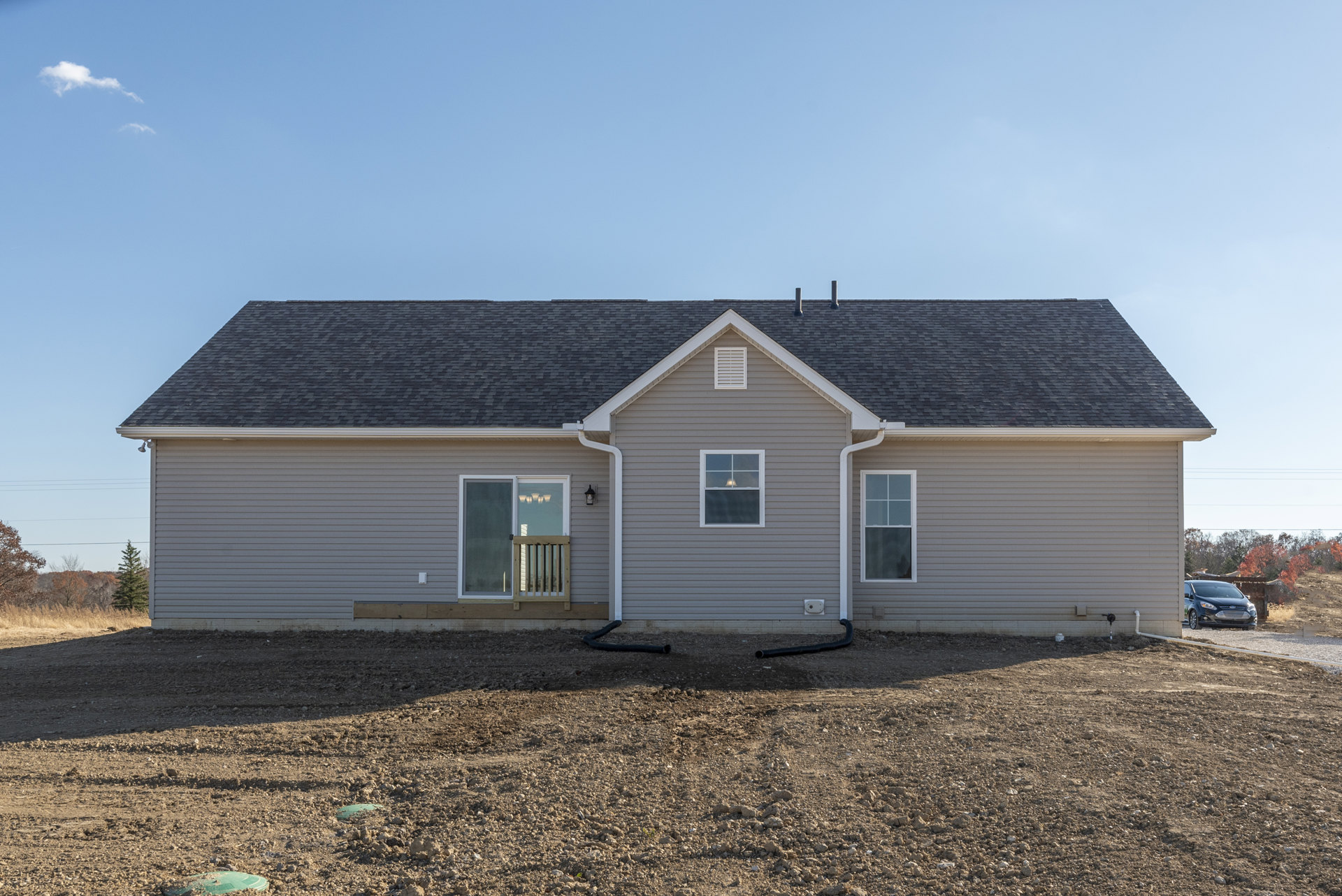 Front exterior of a house with light siding, white-framed windows, wooden railing by the entry door, dirt field in the foreground, black car parked nearby, bell visible in one