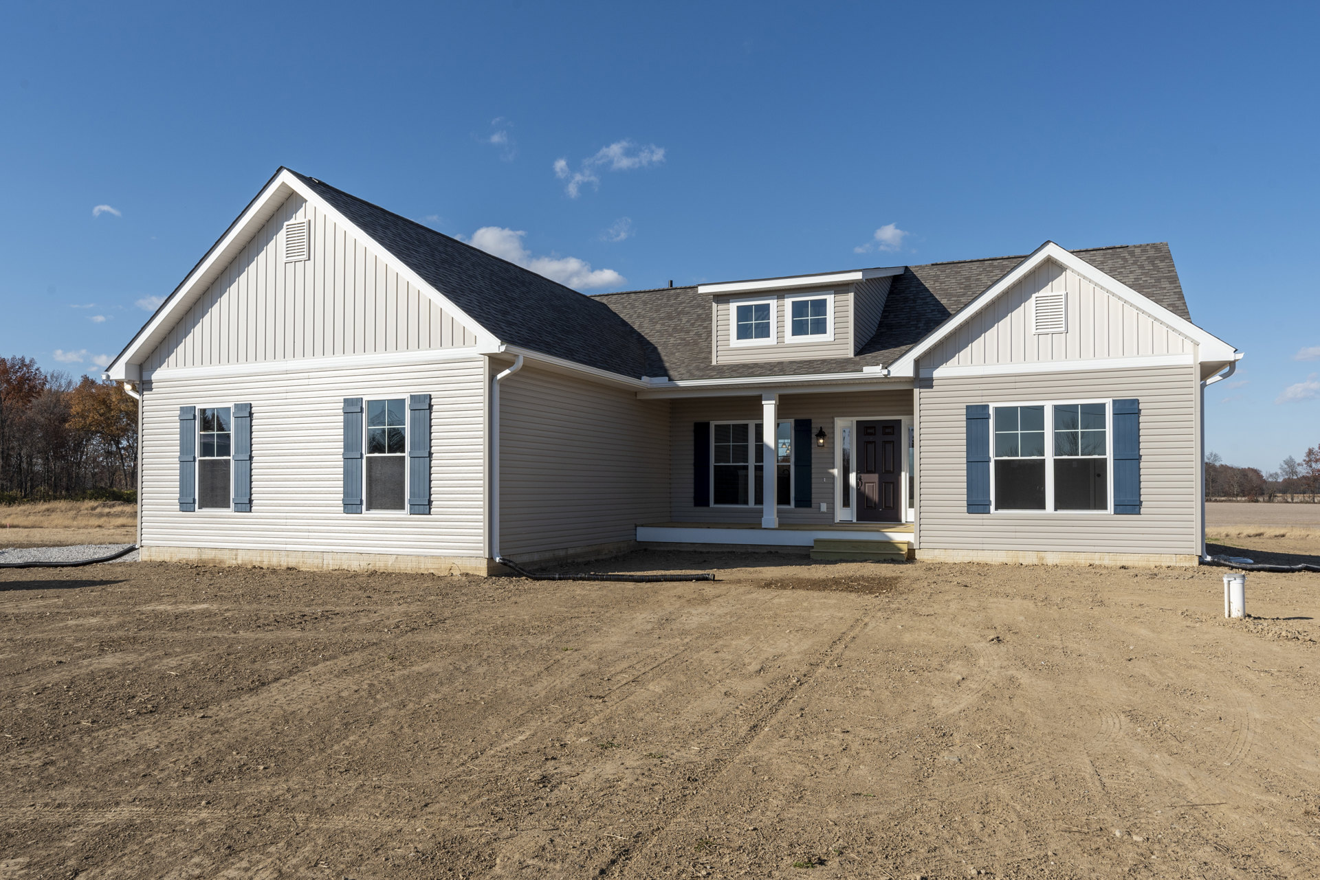 Two-story house with light siding, black front door, white-framed windows, covered porch, and dirt yard with exposed pipe under partly cloudy sky