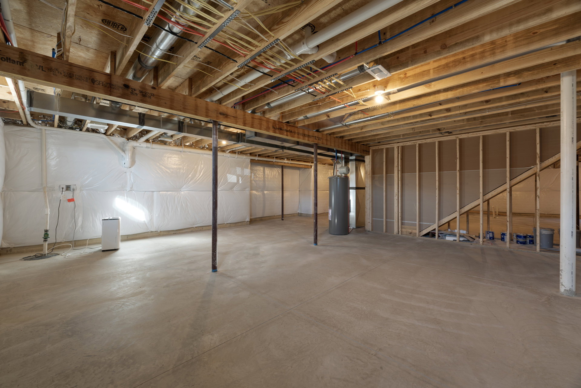 Basement room with exposed wood ceiling beams, concrete floor, white walls, visible steel support pole, pipes, and building insulation.