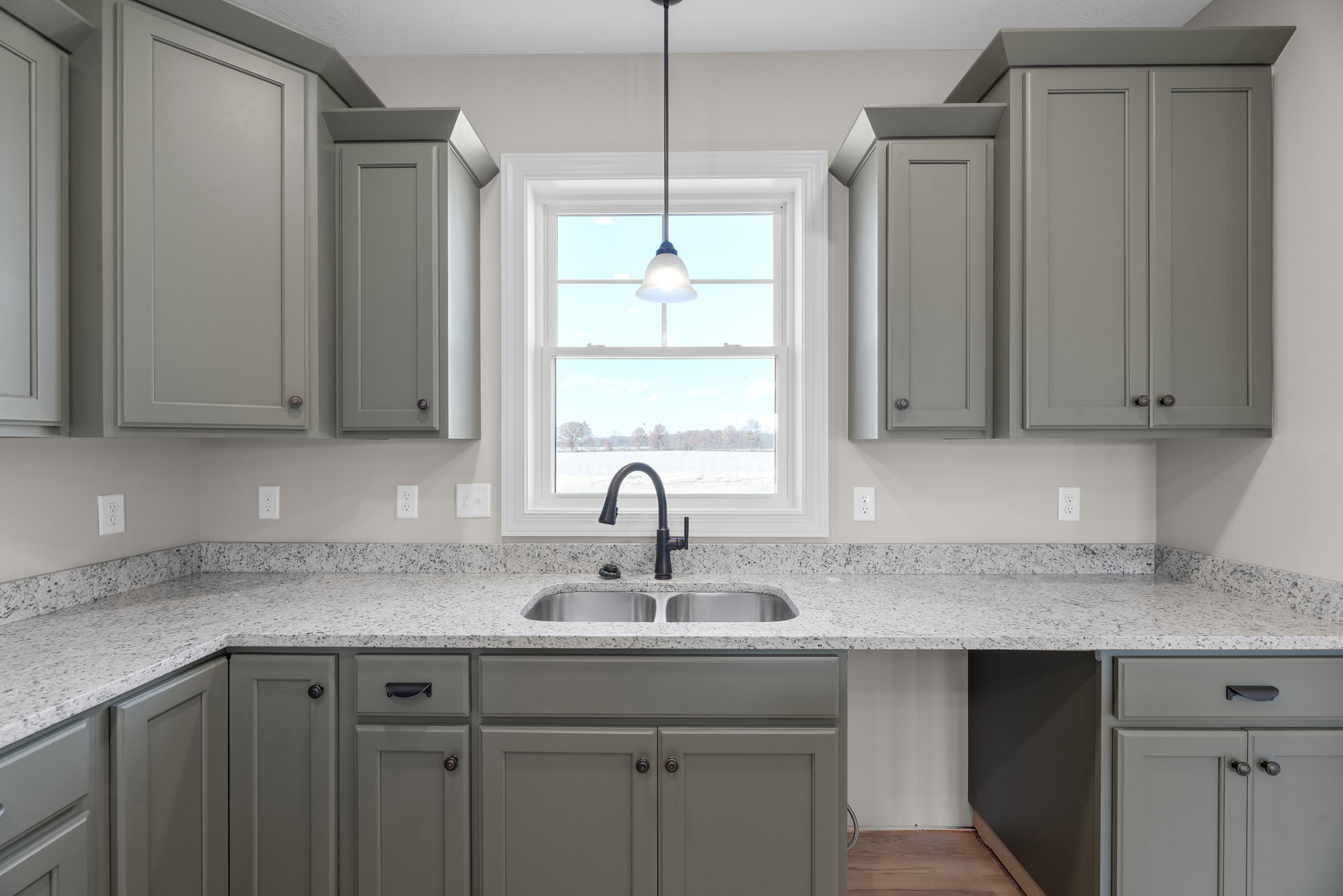 Grey shaker cabinets and white quartz countertops in a kitchen with a stainless steel sink, black faucet, and large window above the sink letting in natural light.
