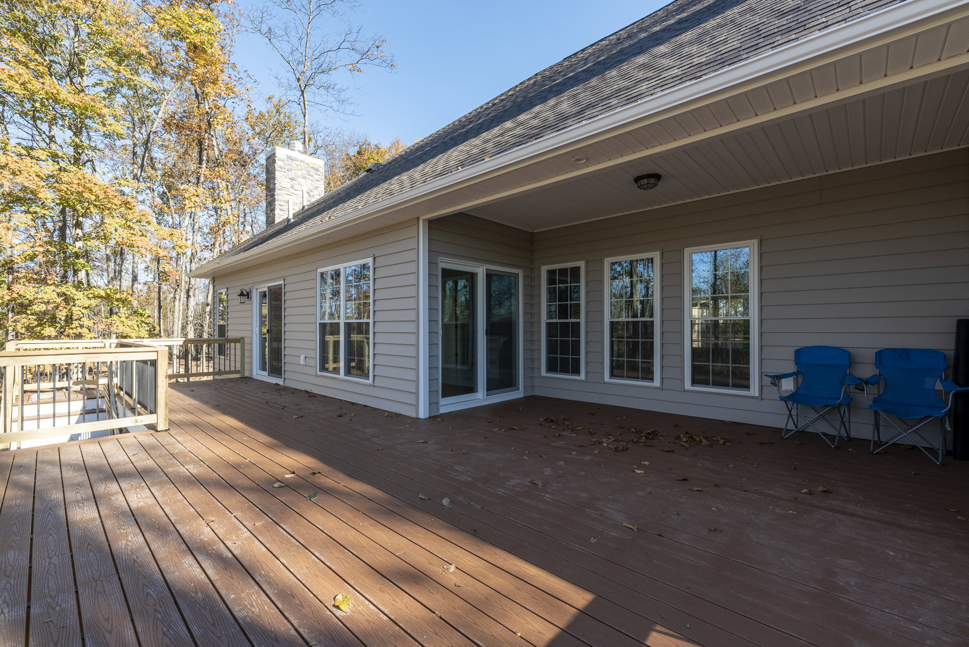 Wood deck with metal railings, blue folding chair positioned against exterior wall, large window with white frame reflecting trees, surrounded by shaded outdoor area and greenery.