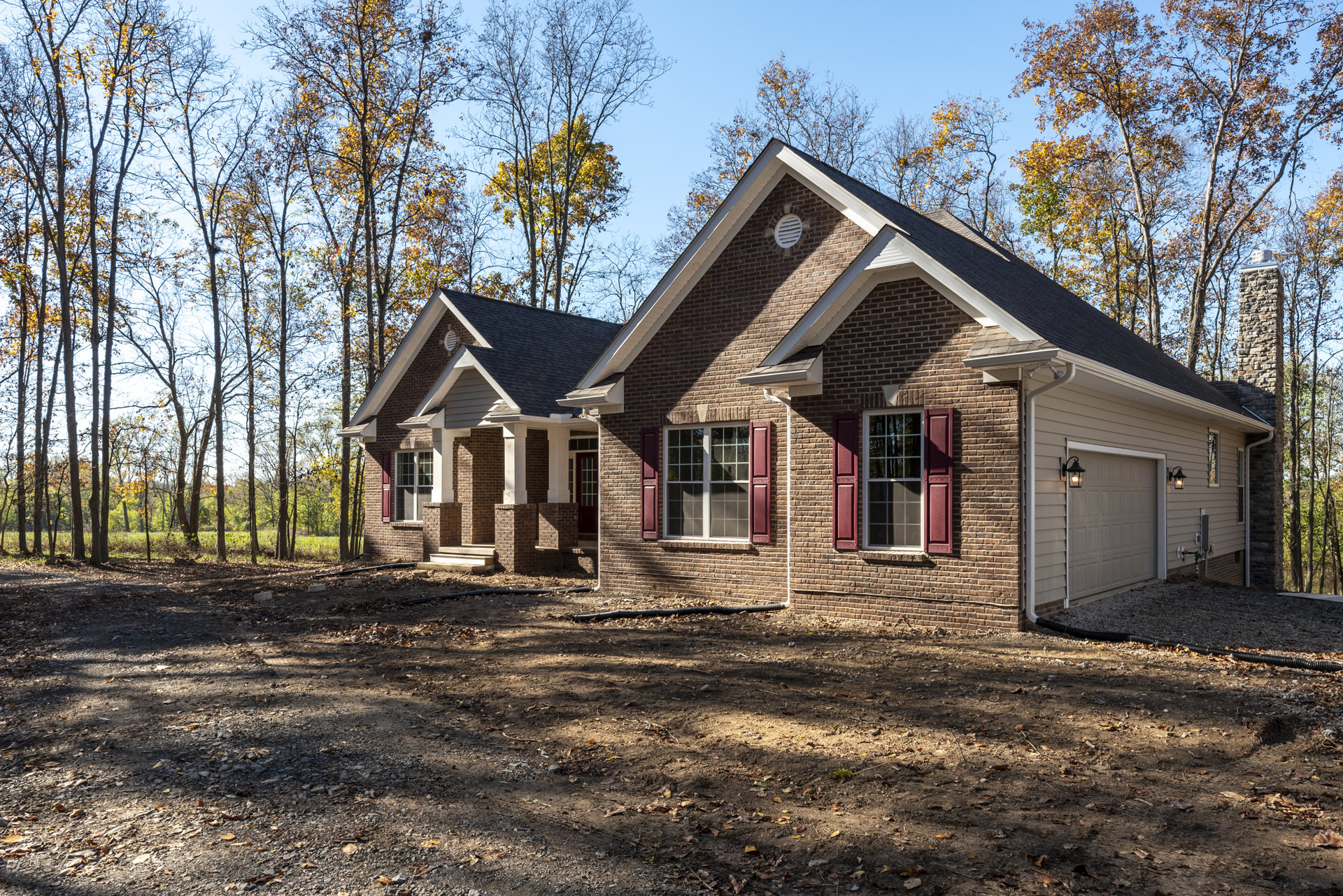 Red brick house with red window shutters, dirt ground in front, garage door with exterior light, mature trees in background.