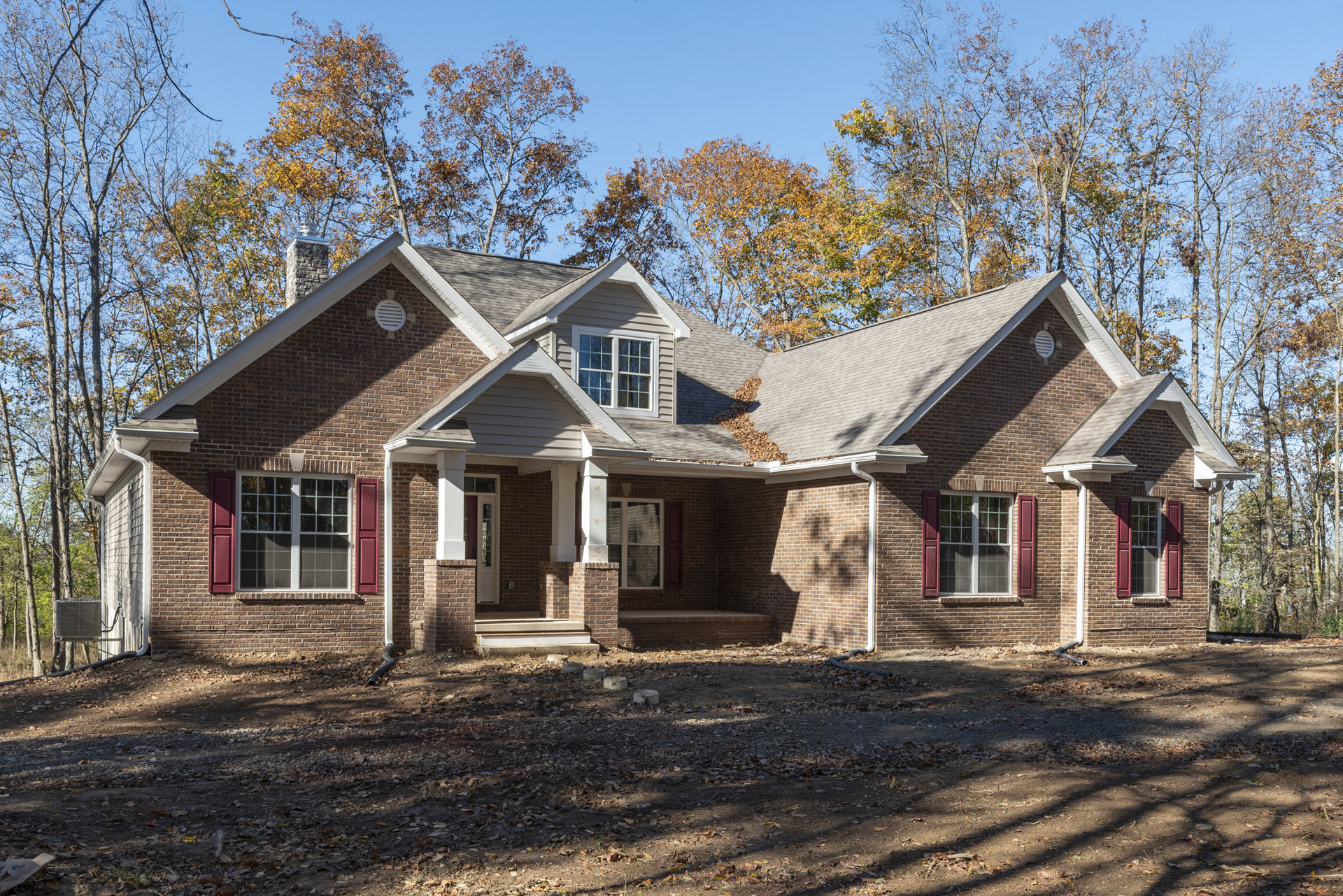 Red brick house with white-trimmed windows and red shutters, surrounded by tall trees; dirt yard with scattered concrete blocks and leaves on the roof.