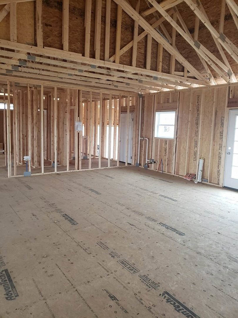 Open room with exposed wood ceiling beams, white-framed window, unfinished plank flooring, and construction materials visible.