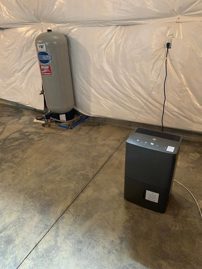 Large grey water heater with red label beside black rectangular control box, both installed on concrete floor with visible pipes and hose connections.