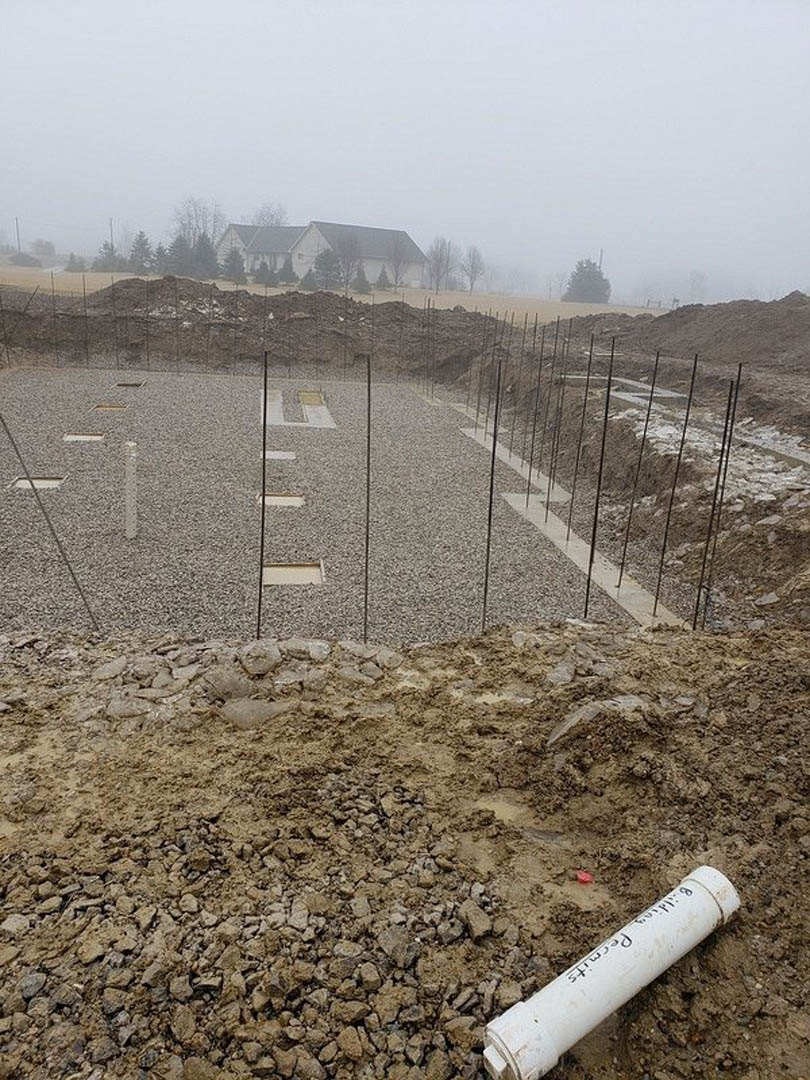 White PVC pipe partially buried in dirt at a residential construction site, piles of soil and gravel road in foreground, house and trees visible in background