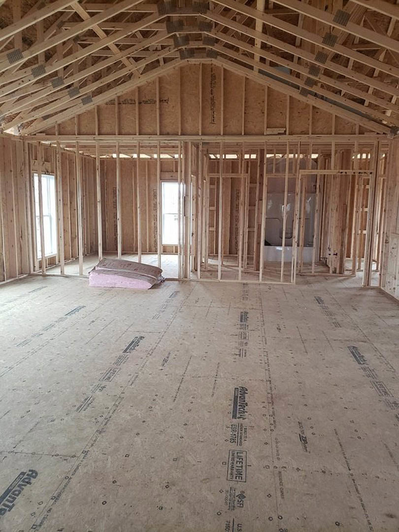 Wood-framed room under construction with exposed ceiling trusses, unfinished floor, stacked bricks, and a window with a white frame.