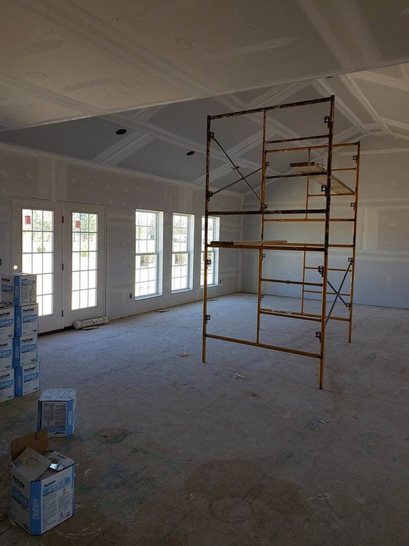 Scaffolding set up in unfinished room with white double doors, several windows with white frames, and stacked cardboard boxes on bare floor.