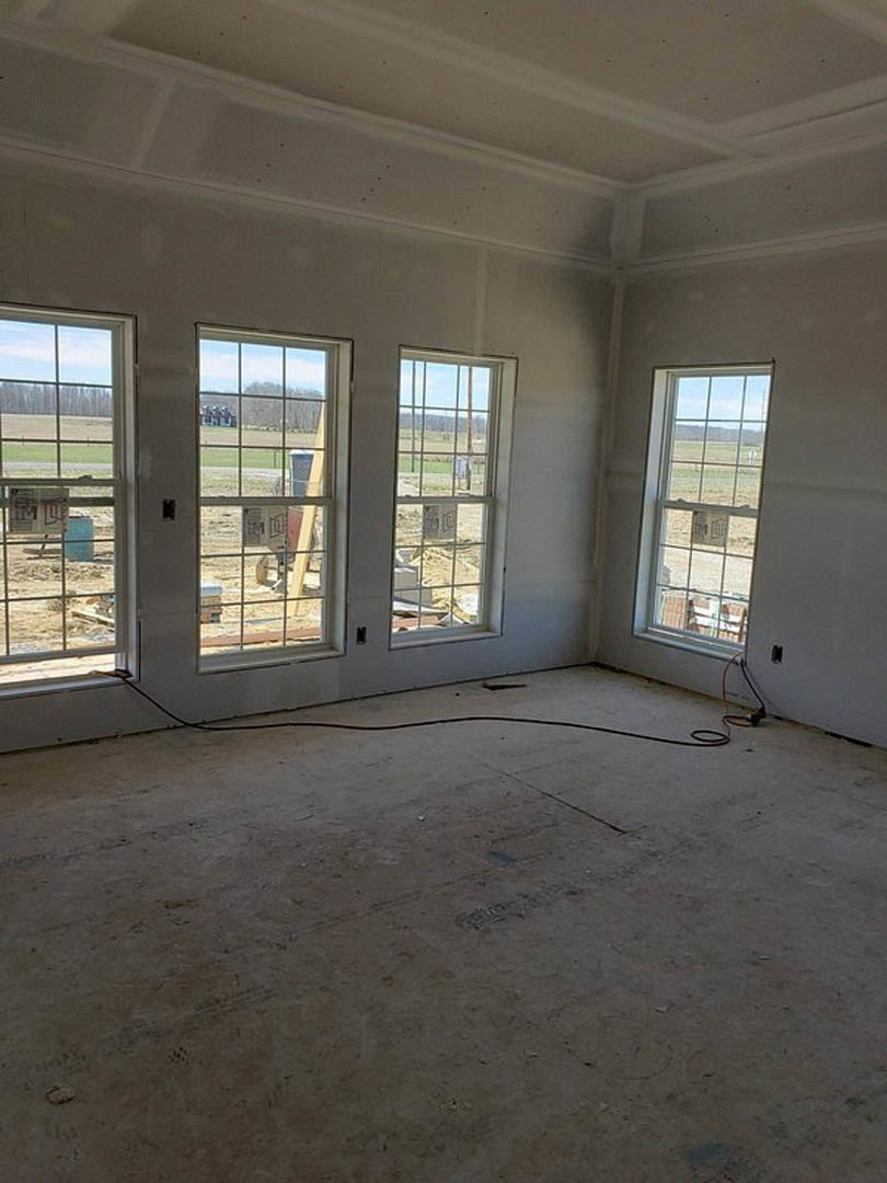 Spacious unfinished room featuring large windows, exposed wire on concrete floor, partial view of open field and nearby building, natural daylight illuminating construction