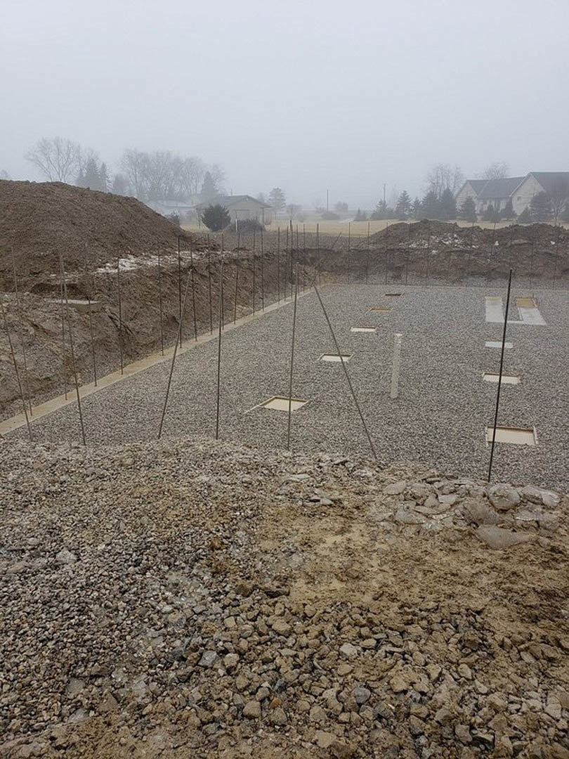 Construction site with several square foundation holes in dirt, scattered rocks, and trees in the background under a foggy sky