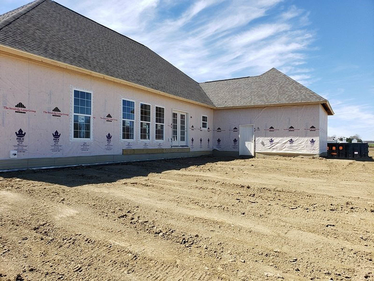 Framed house under construction on a dirt lot, white exterior door and window with sign, cloudy sky overhead
