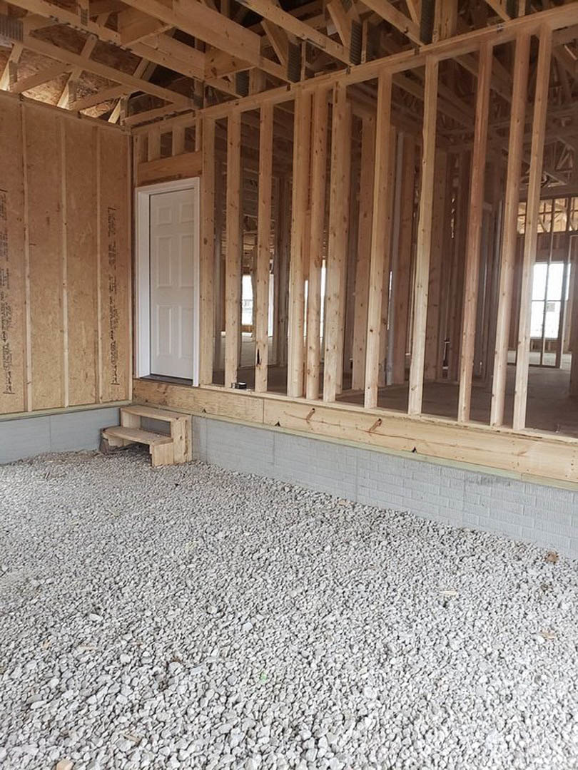 White door with wooden frame set in a stone wall, wooden bench along the wall, white gravel floor, exposed wood beams and planks, unfinished interior of a house under construction.