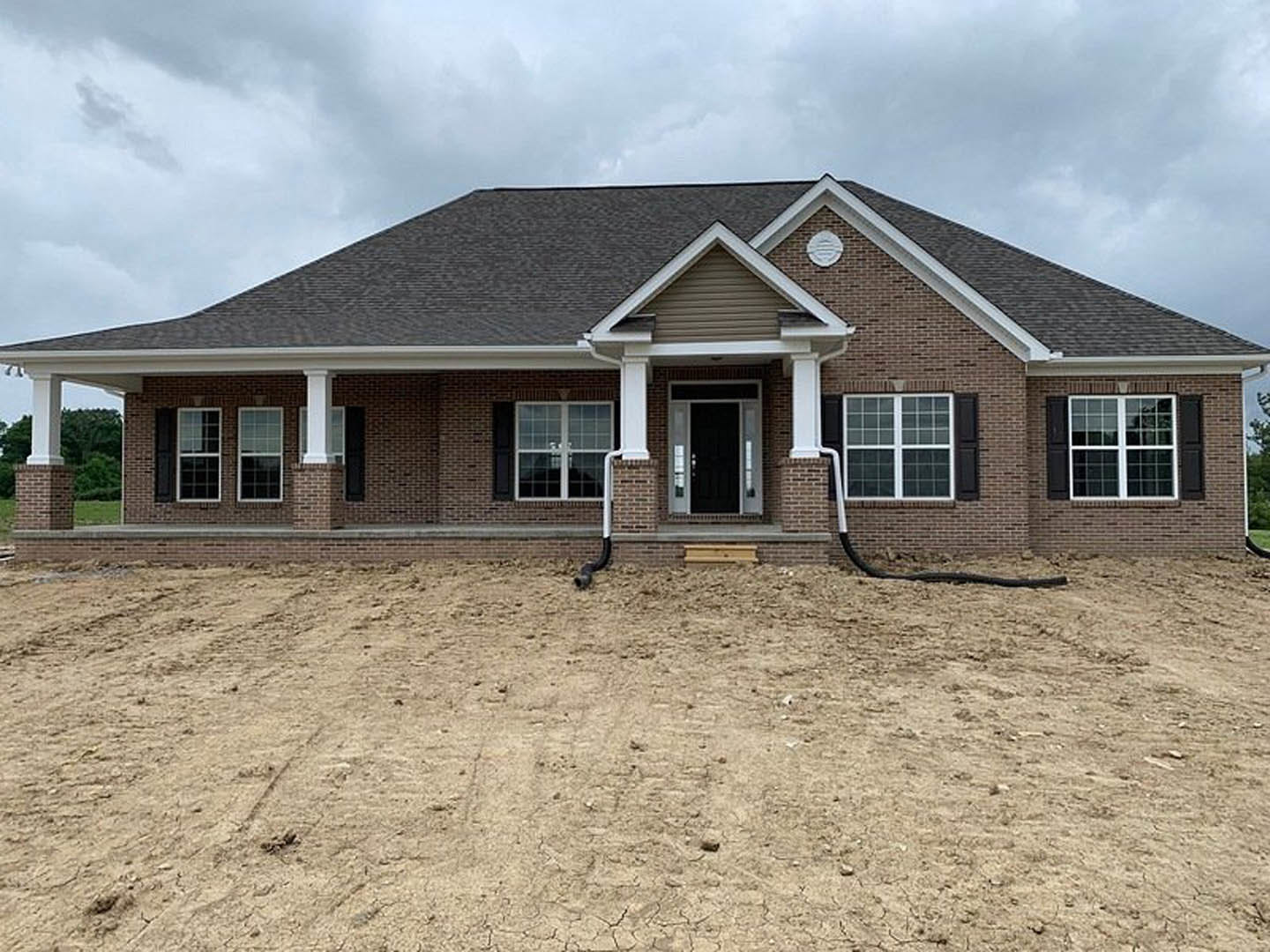 Brick house under construction with exposed dirt yard, black glass-paneled front door, white-framed windows, and cloudy sky overhead