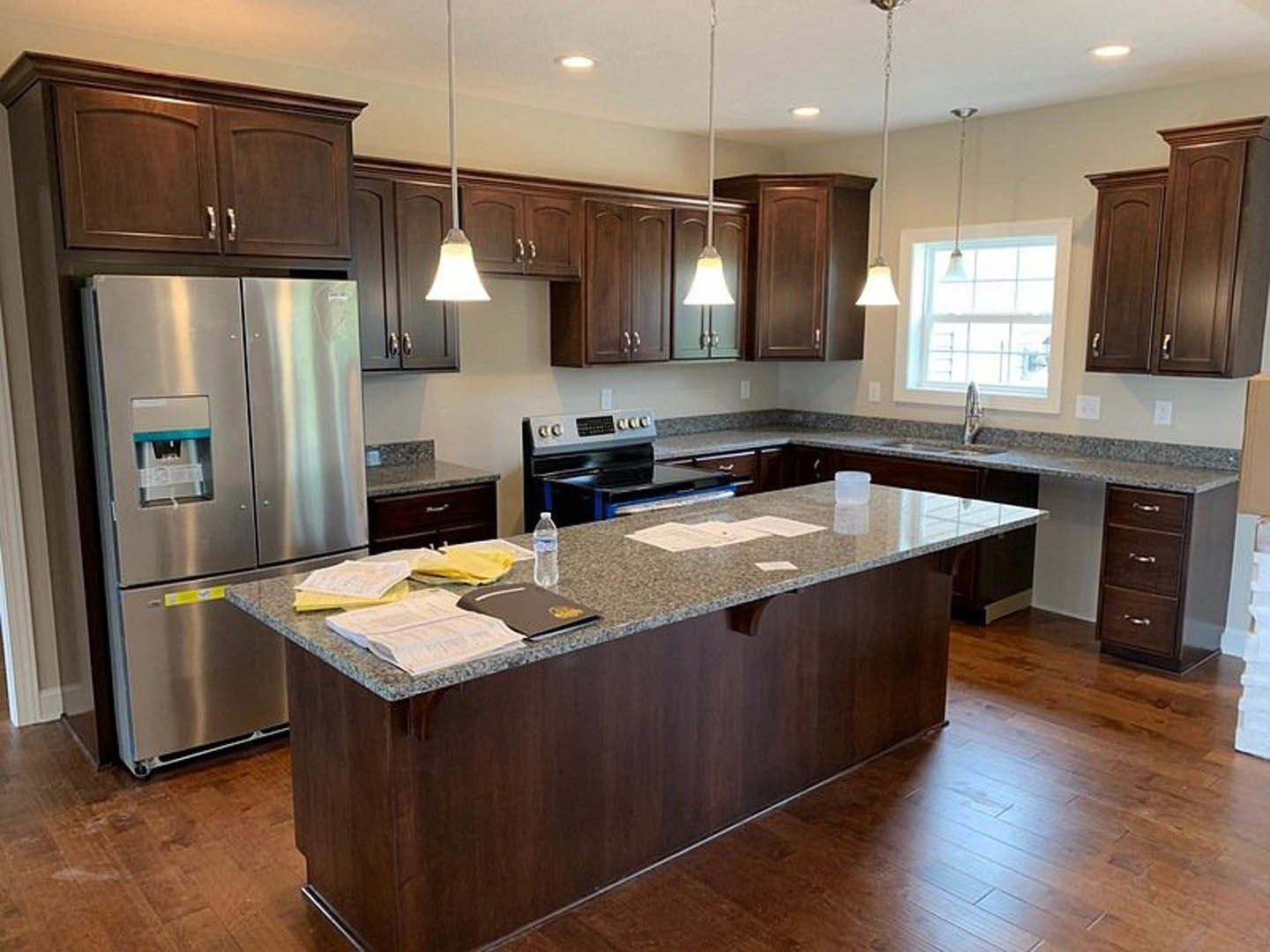 Spacious kitchen featuring a large quartz island, stainless steel refrigerator and stove, white cabinetry, wide window above a farmhouse sink with chrome faucet, pendant lamp