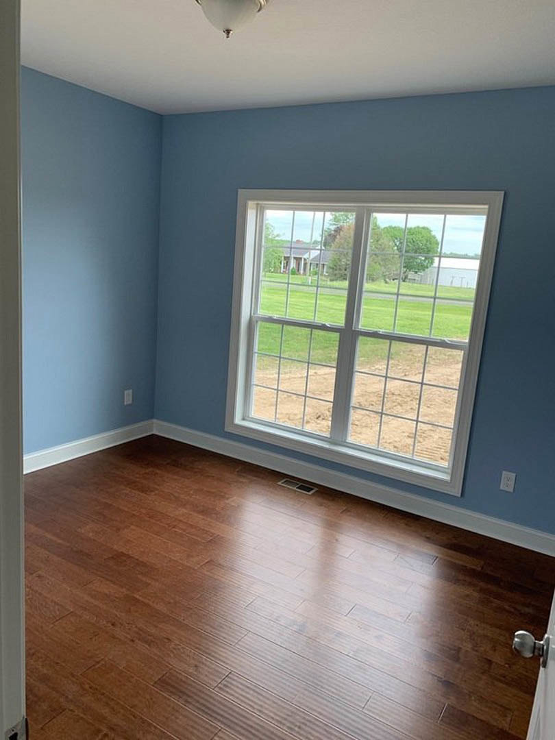 Sunlit room featuring a large window overlooking green grass, hardwood floors, white walls, and a modern ceiling light fixture
