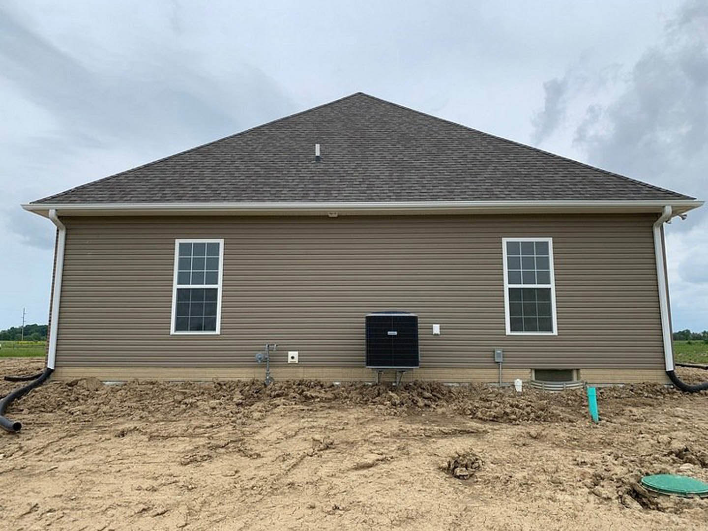 Partially built home with white framed window, black square vent, gray shingle roof, and light siding, surrounded by dirt and construction materials under a cloudy sky