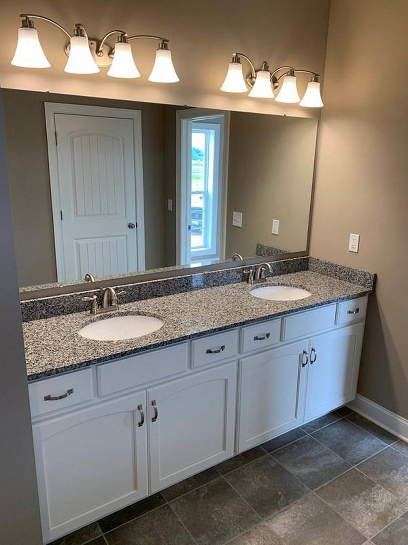 Bathroom with expansive mirror above white vanity featuring black speckled countertop, dual-lamp light fixture, white door with silver handle, and window with clear glass pane.
