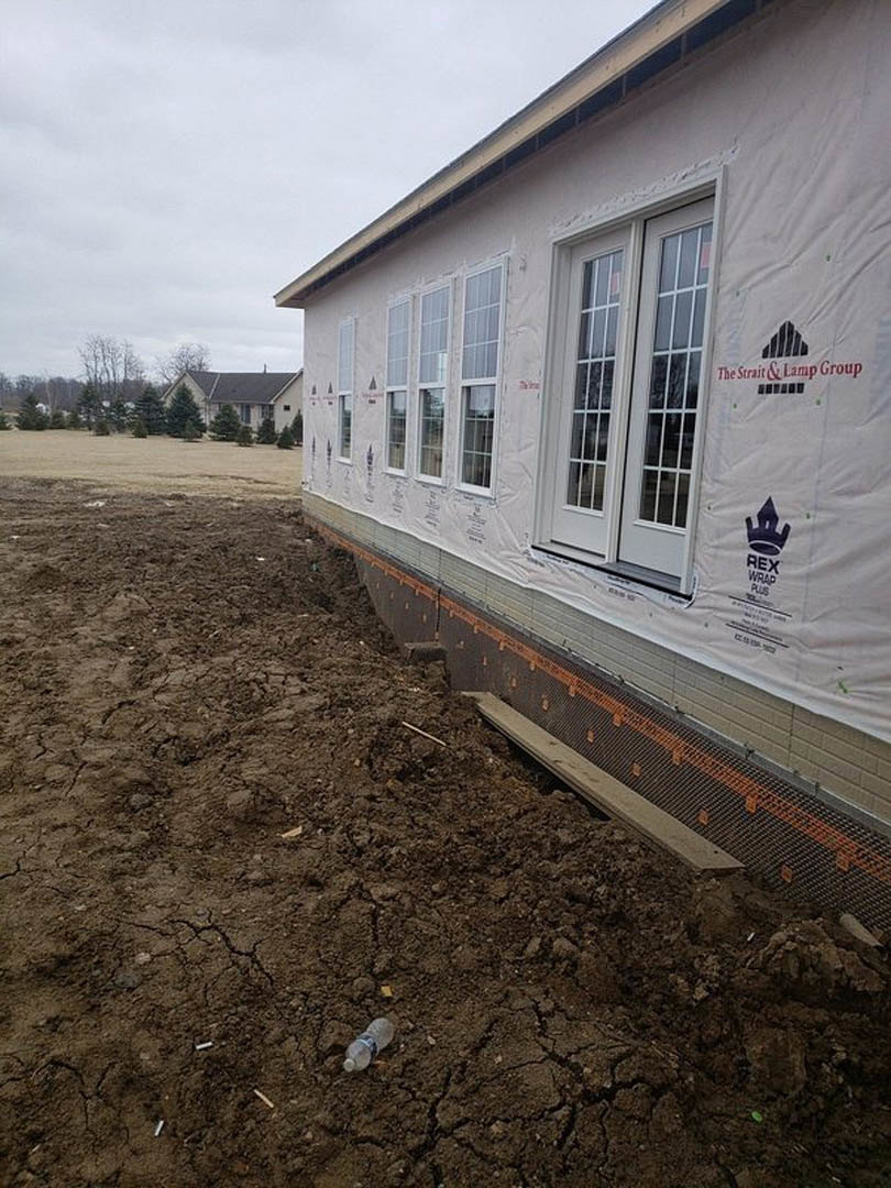 Modern house under construction with white protective covering, exposed dirt ground, plastic bottle, large windows, and trees in the background