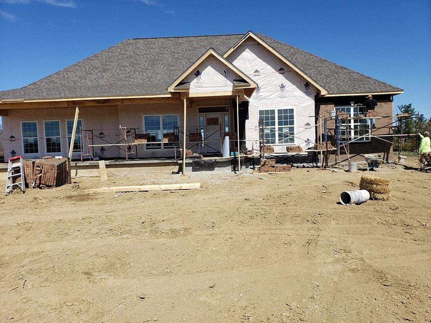 Partially built brick house with exposed framing, dirt field in foreground, temporary fence, ladder leaning against exterior wall, visible white door with glass panes, scattered