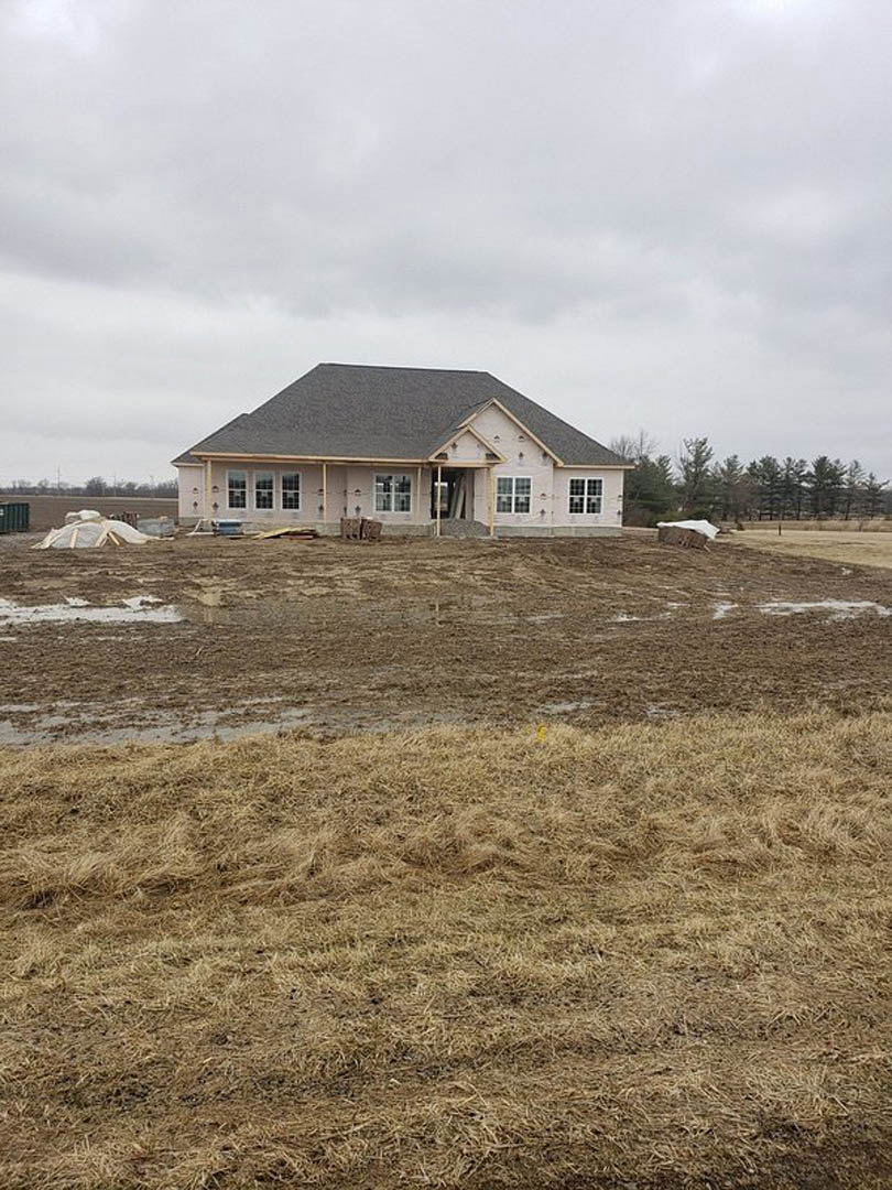 Wood-framed house under construction with shingled roof, surrounded by grassy field, white construction bags near perimeter, wire fence, and trees in background under partly cloudy
