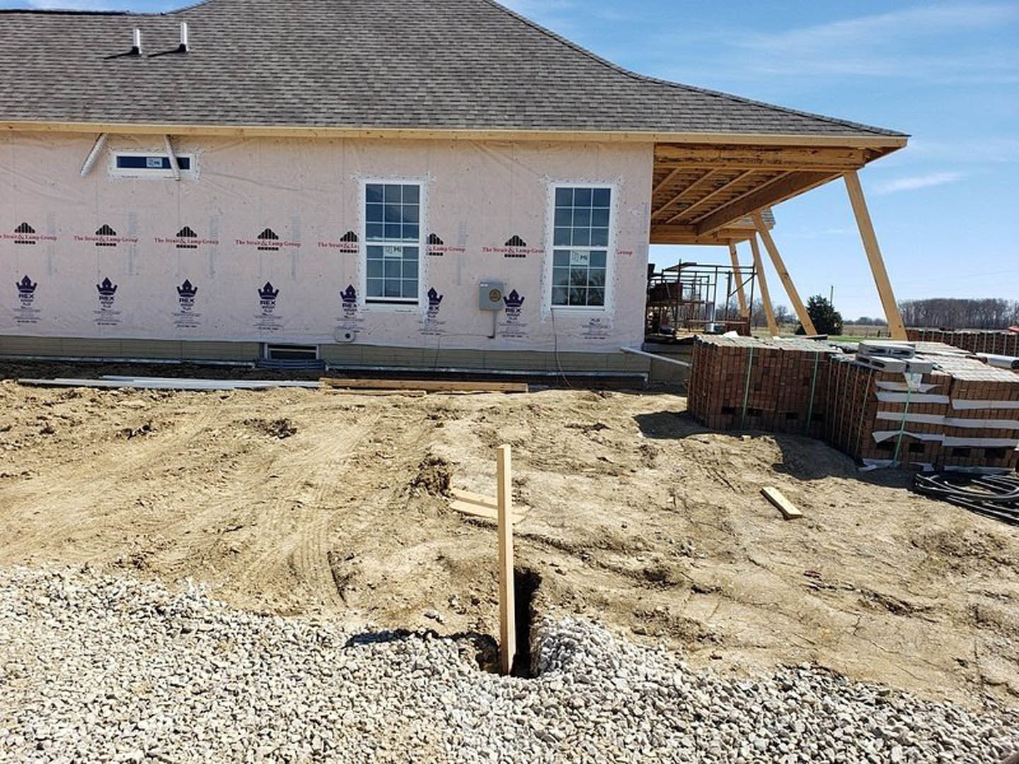 Partially built house with exposed framing, installed windows, stack of bricks, wooden post set in a dirt hole, construction materials scattered on bare ground