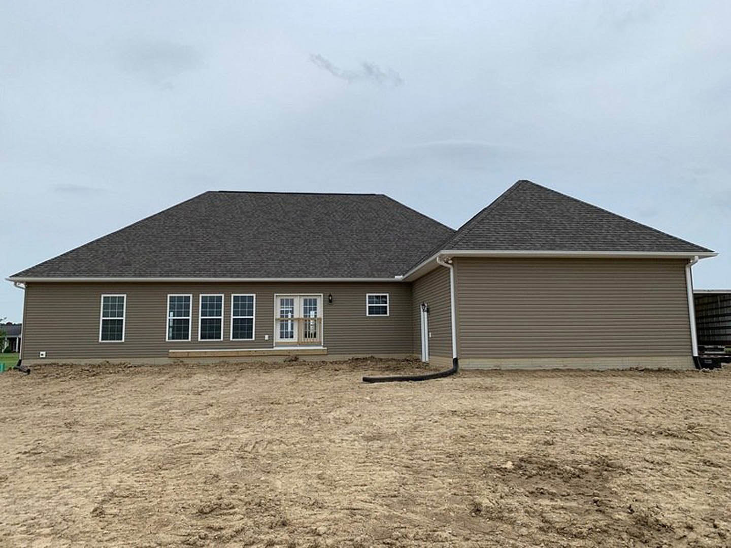 Partially built house with gray siding, white-trimmed windows, shingled roof, and dirt yard with garden hose under cloudy sky