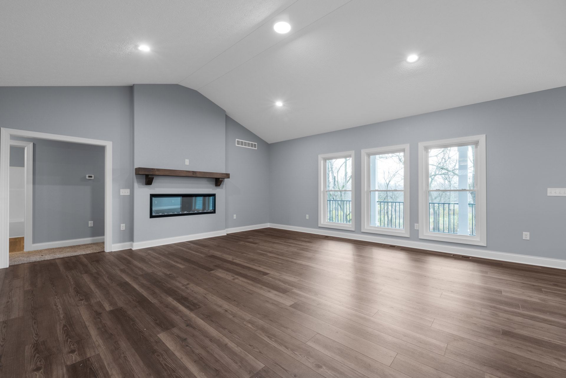 Spacious living area with hardwood flooring, modern fireplace, ceiling light fixture, glass wall with black frame, window overlooking trees, and wood accent board on plaster wall