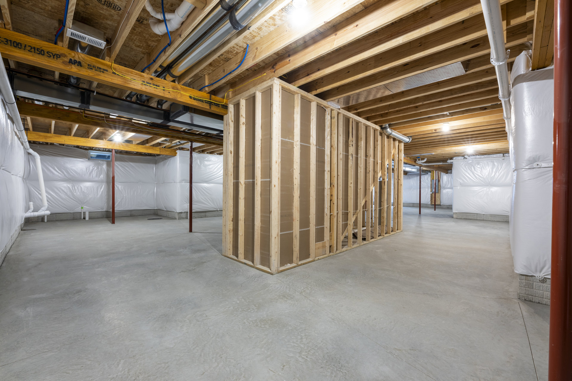 Exposed wood framing and beams with visible ceiling pipes, white plastic wall covering, unfinished floor, and building insulation in a residential interior.
