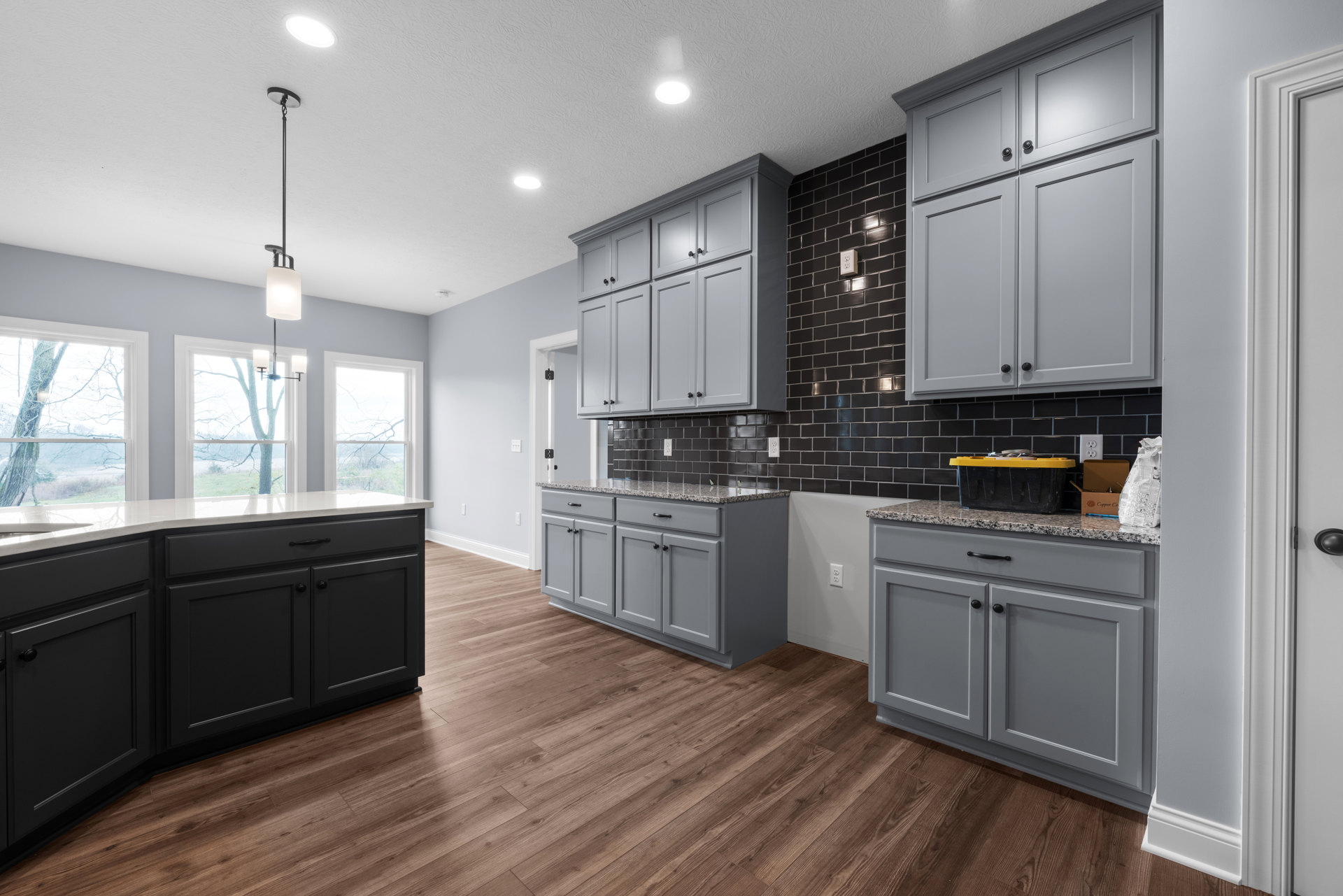 Grey kitchen cabinets with matte black tile backsplash, stainless steel sink, white countertops, and a window showing snowy trees outside