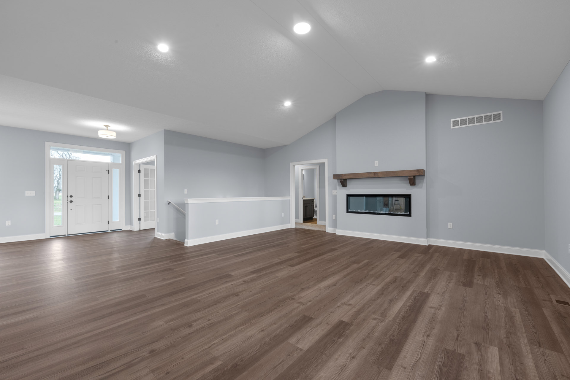 Spacious room with hardwood flooring, central fireplace with black surround, white walls, ceiling light fixture, white door with glass window, wall vent