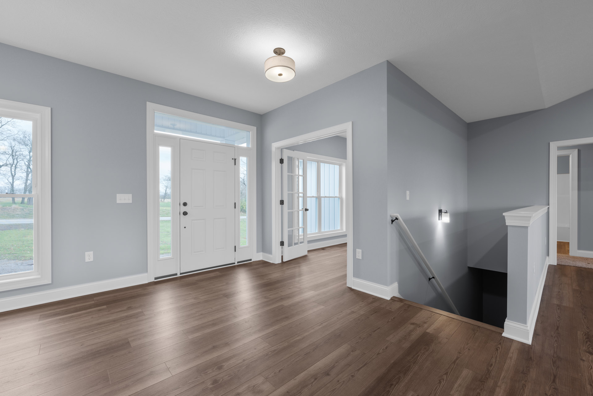 Hallway with wood flooring, white walls, staircase with white railing, white door featuring glass panes, and ceiling light fixture with white shade