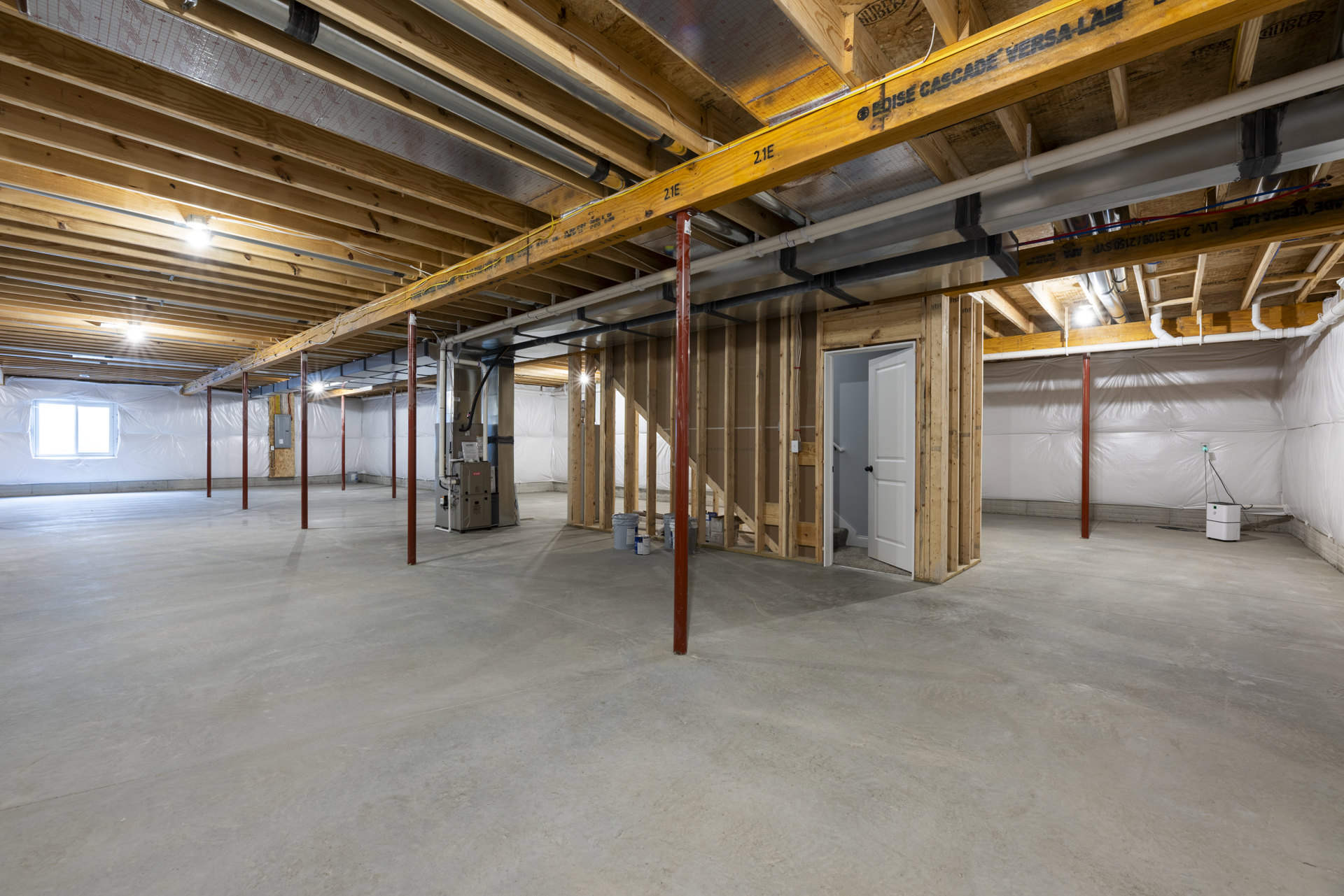 Basement room with exposed wooden ceiling beam featuring black writing, concrete floor, red support pole, white window cover, white door with handle, and white electrical box with
