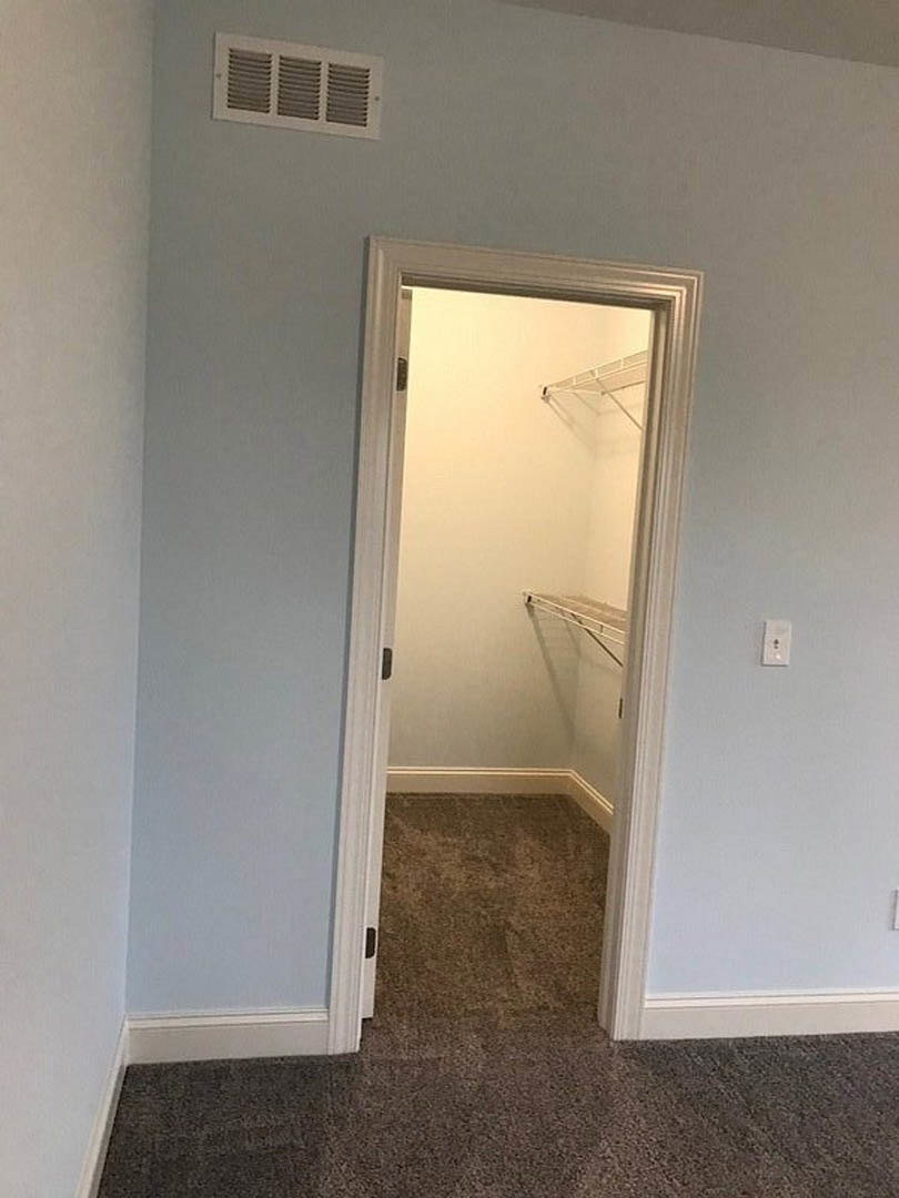 Open closet door revealing metal rack shelving, carpeted floor, wall vent, and white plaster walls in a residential bedroom.
