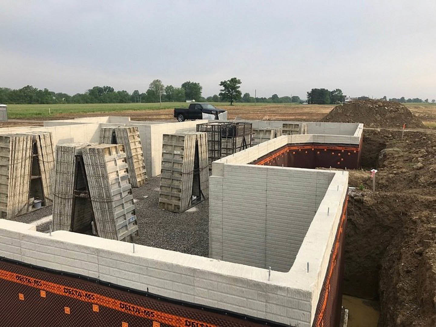 Construction site with stacked concrete blocks and slabs, black truck with open door parked in background, tall unfinished building, trees visible in distance under open sky
