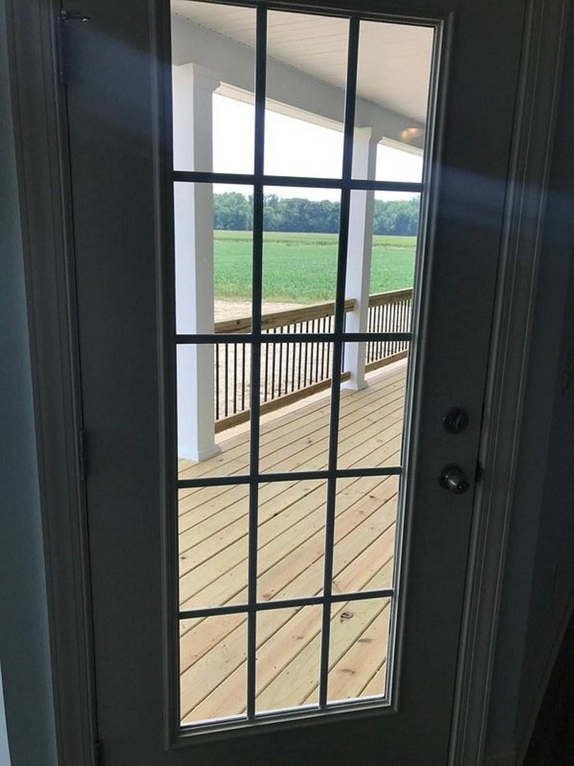 Glass door framed in black iron opens to a wood deck overlooking a grassy field, white interior walls with black trim visible