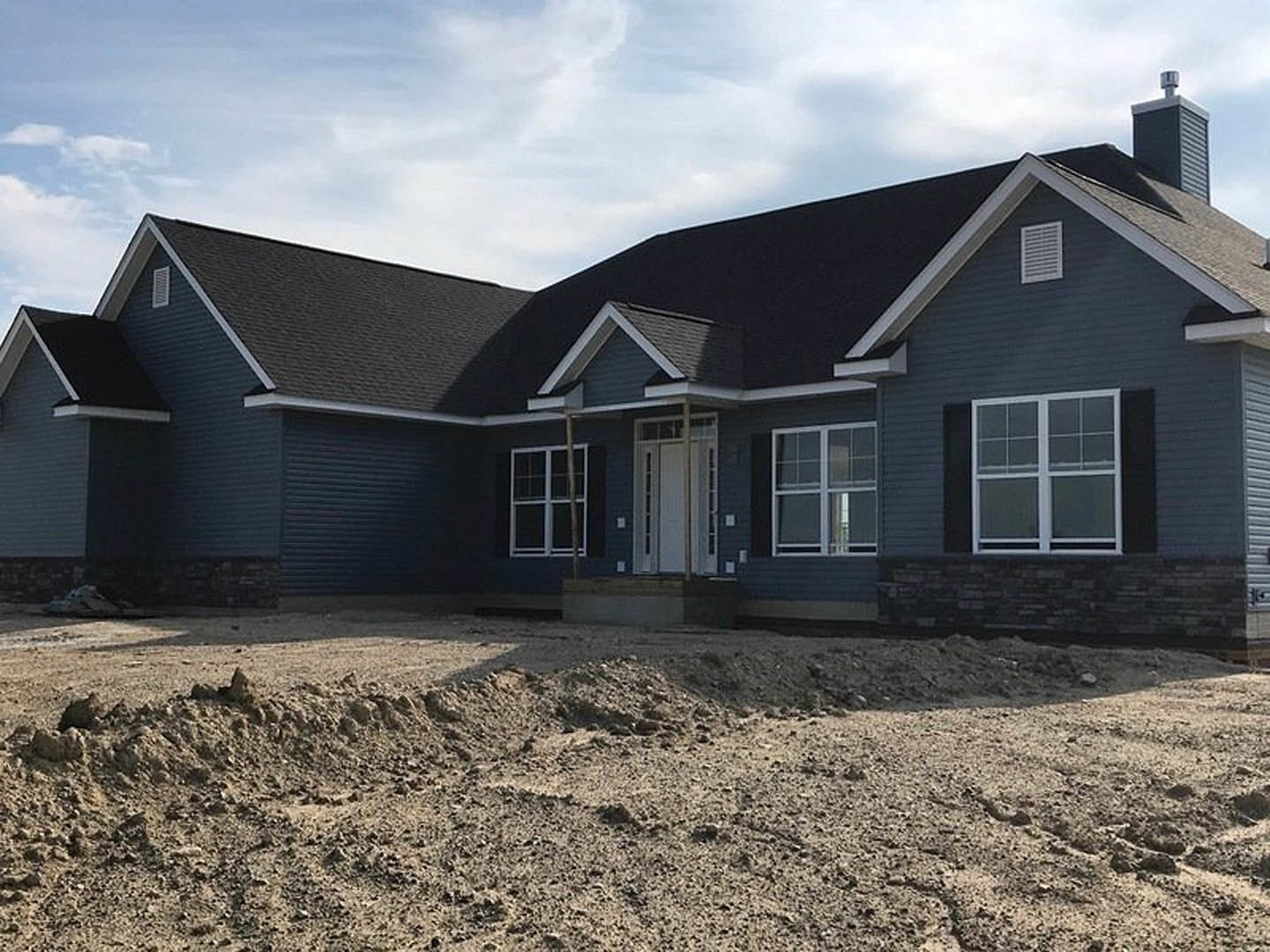 Two-story house under construction with white framed windows, glass-paneled door, covered porch, and piles of dirt in the foreground