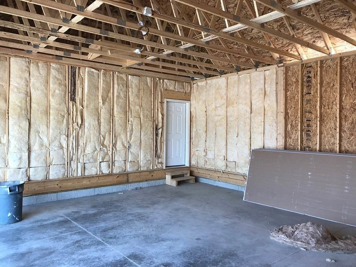 White interior room featuring two white doors with brown frames, light plaster walls, exposed ceiling beams, and a concrete floor with a white border; bench and trash can visible