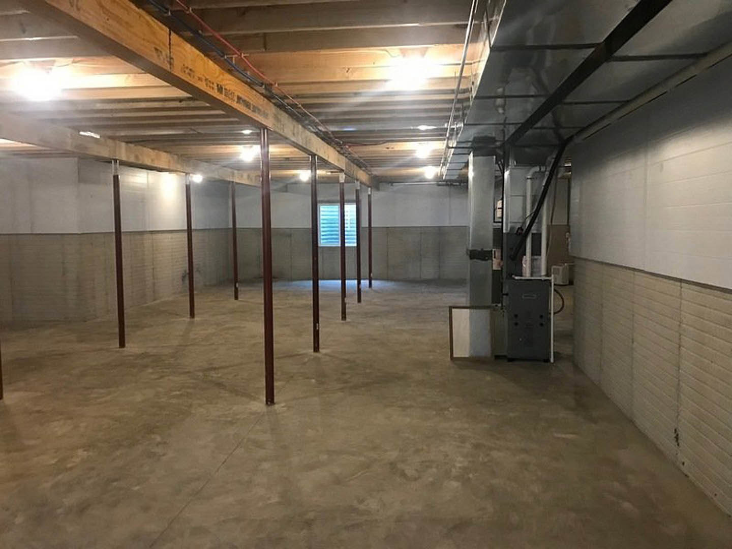 Concrete-floored room with exposed metal pole, wood ceiling beam fitted with recessed lights, window covered by blinds, and black rectangular vent with holes on wall