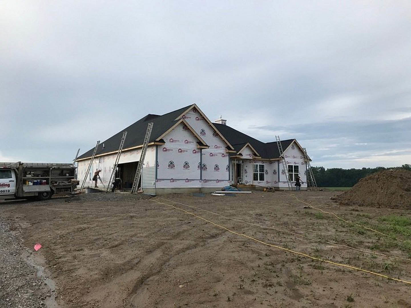 Framed house under construction with exposed wood, ladders leaning against exterior, large pile of dirt in foreground, construction truck parked nearby, cloudy sky overhead