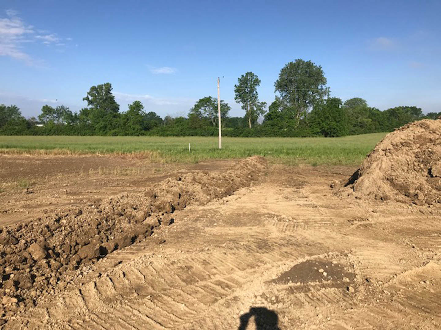 Dirt road bordered by a grassy field, pile of soil in foreground, leafy trees and utility pole under a partly cloudy blue sky