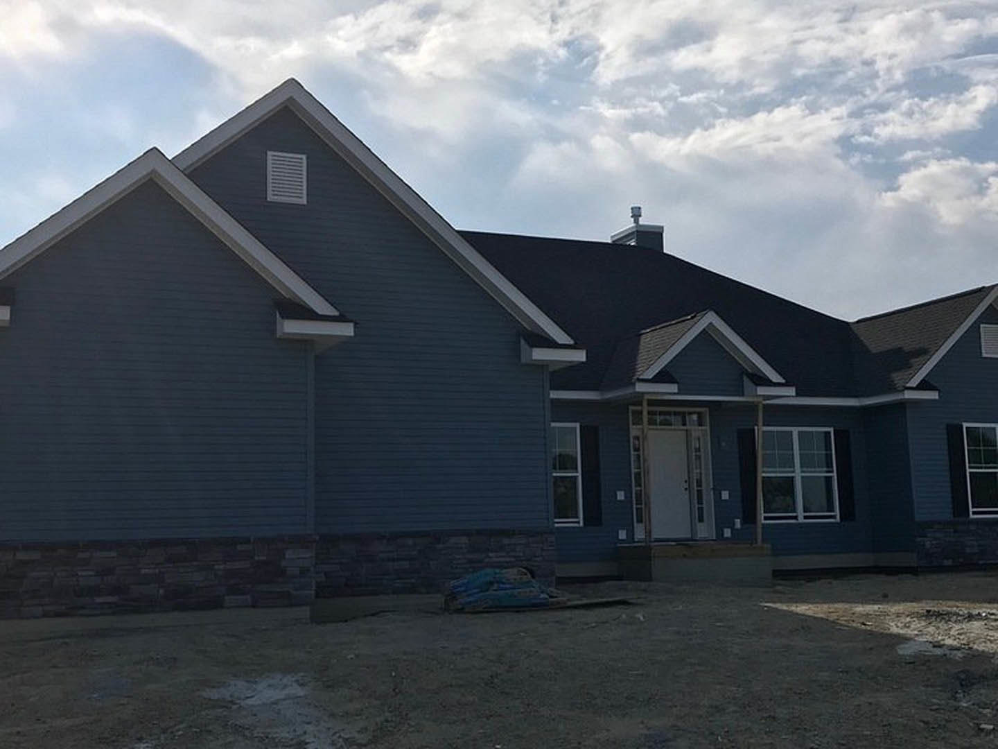 Partially finished house with white framed window, white glass panel door, exterior wall vent, exposed siding, and dirt yard under blue sky with scattered clouds
